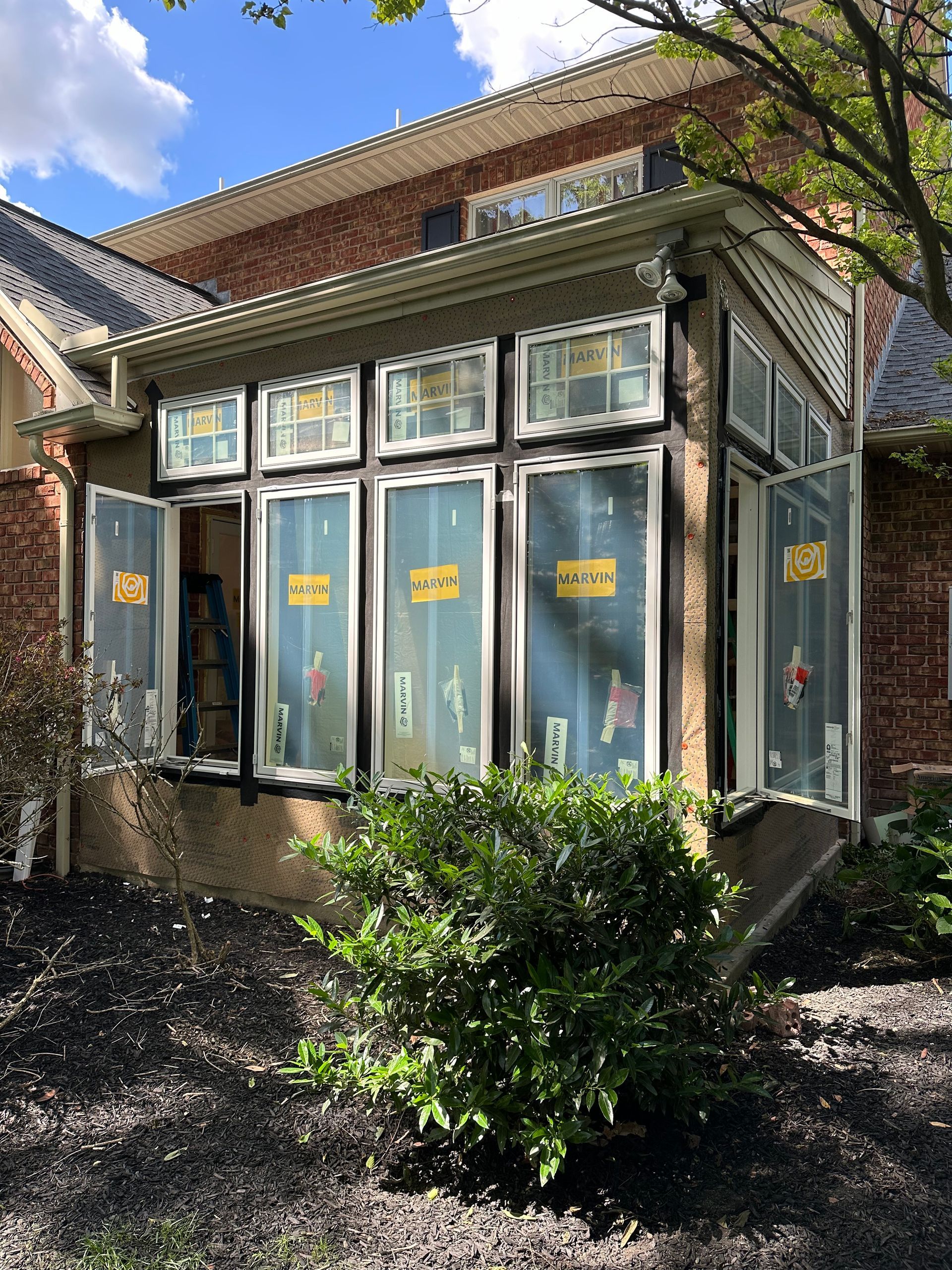 Building with large windows, some open, with yellow signs. Black mulch and green bush in front.