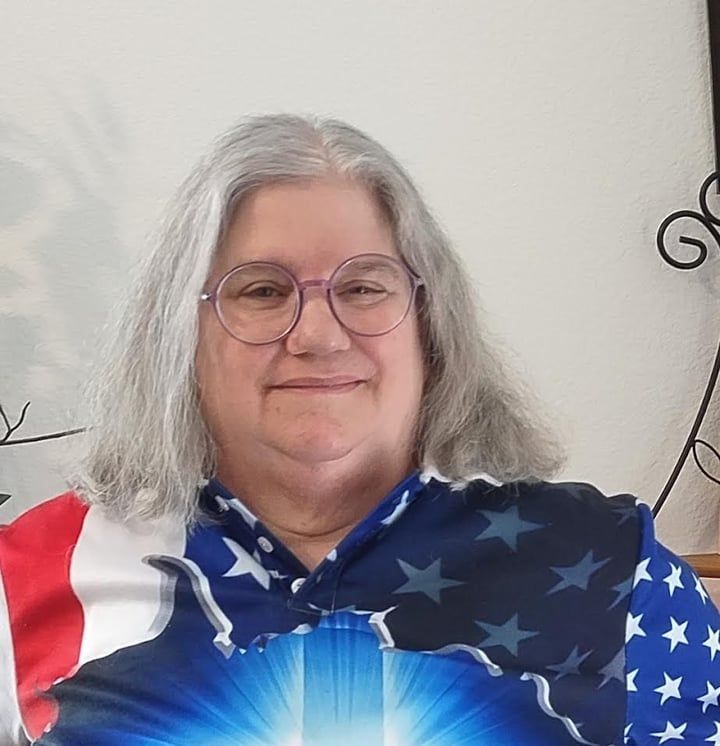 Person in glasses wearing an American flag shirt, smiling against a light-colored wall.