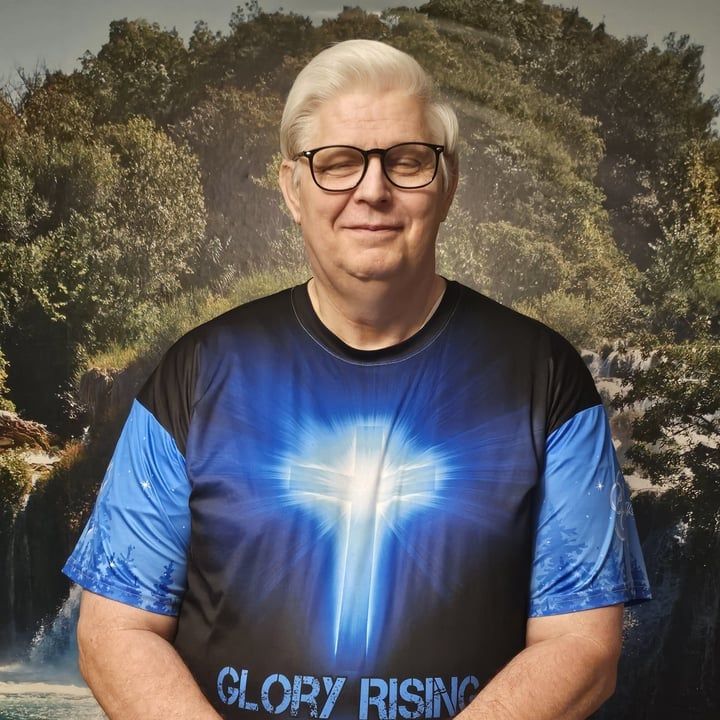 Man in glasses wearing a blue “Glory Rising” T-shirt, standing before a painted forest backdrop.