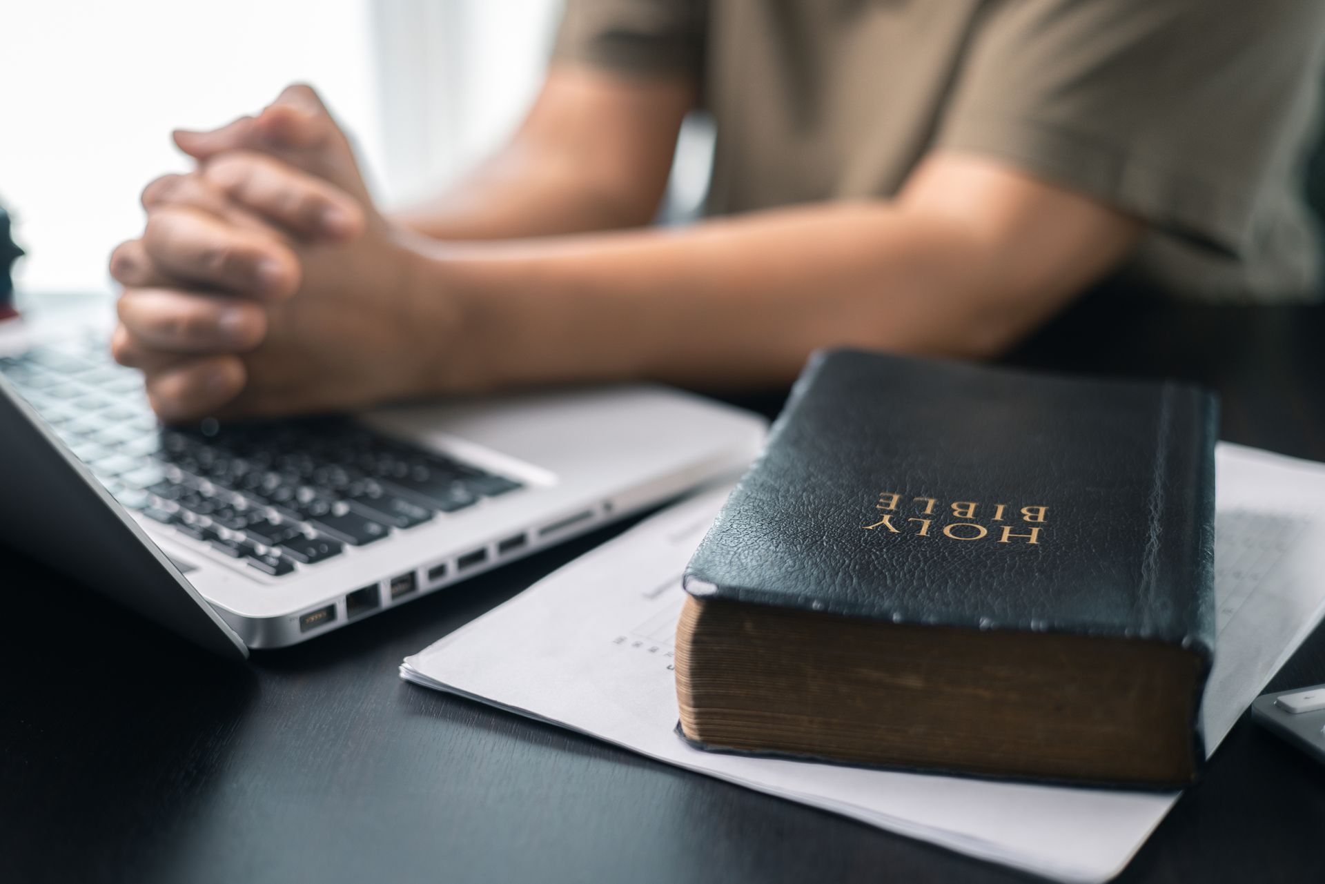 Hands clasped beside a laptop and Bible on a desk, suggesting study or prayer
