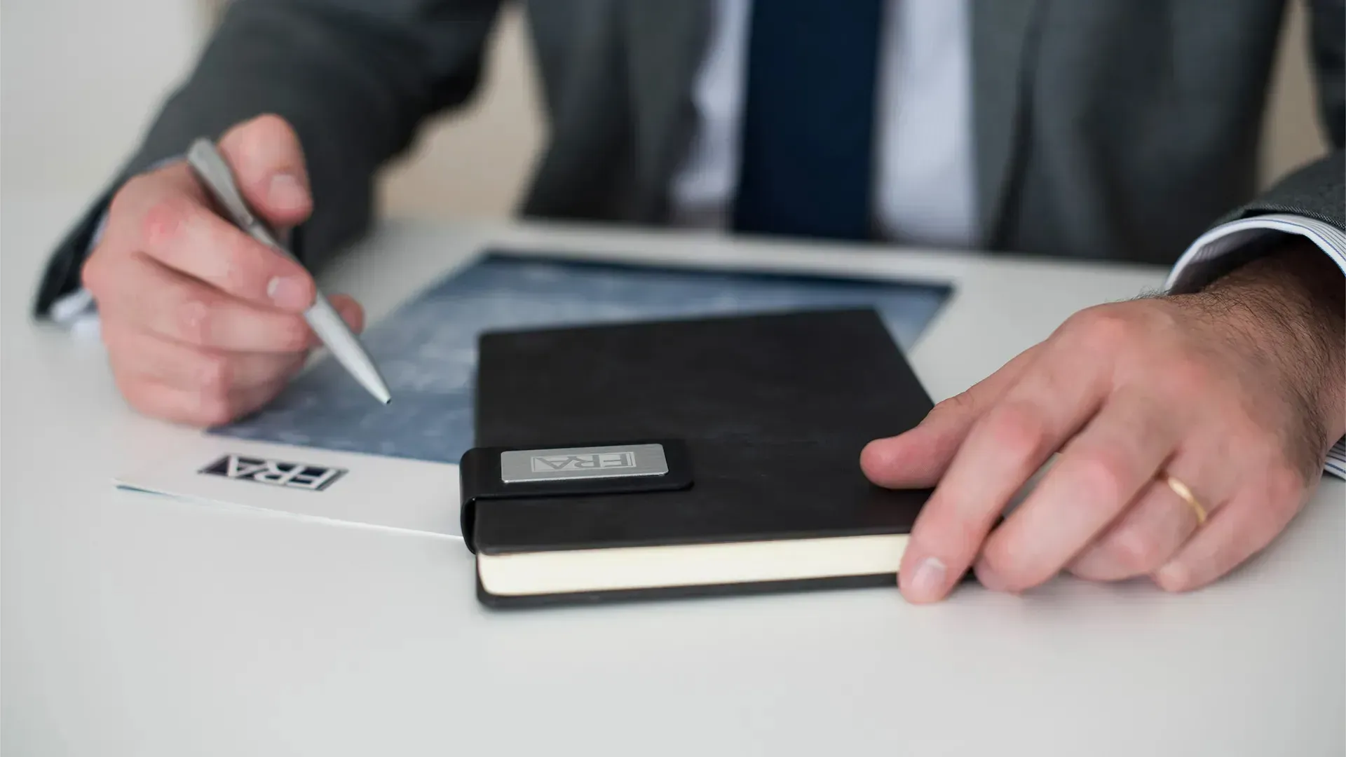 A person in a suit sits at a desk with a pen in hand, resting their other hand on a closed black notebook.