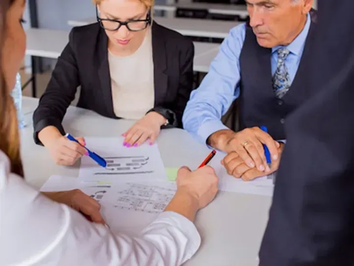 Three business professionals in formal attire sit at a table reviewing documents with pens in an office setting.