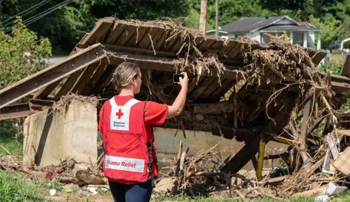 A Red Cross volunteer in a red vest records damage at a site covered in debris from a flood.