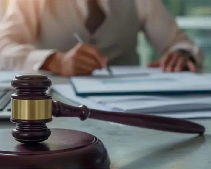 A wooden gavel resting on a block in the foreground with a blurred person writing on documents in the background.