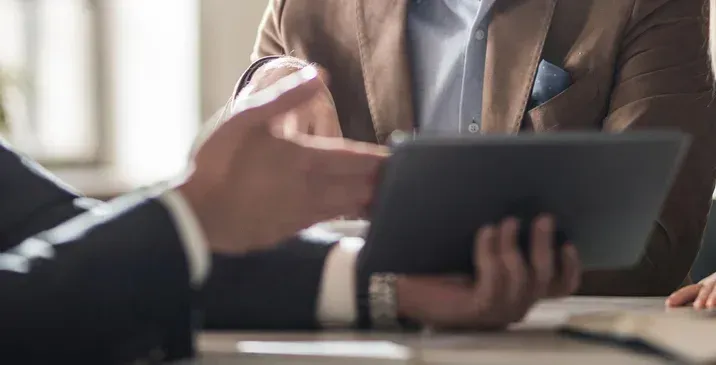 Two professionals in business attire sitting at a desk and reviewing information on a tablet.