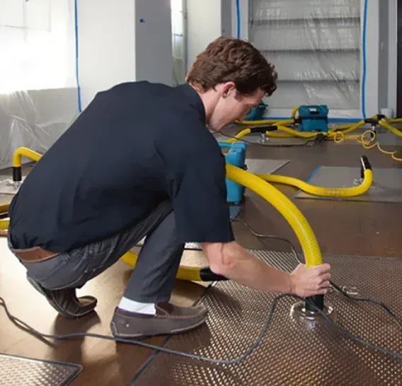 A technician crouches on a wooden floor, connecting yellow hoses to metal panels as part of a professional water extraction.