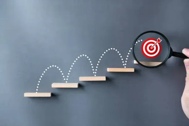A magnifying glass focuses on a red target icon atop a series of ascending wooden blocks against a grey background.