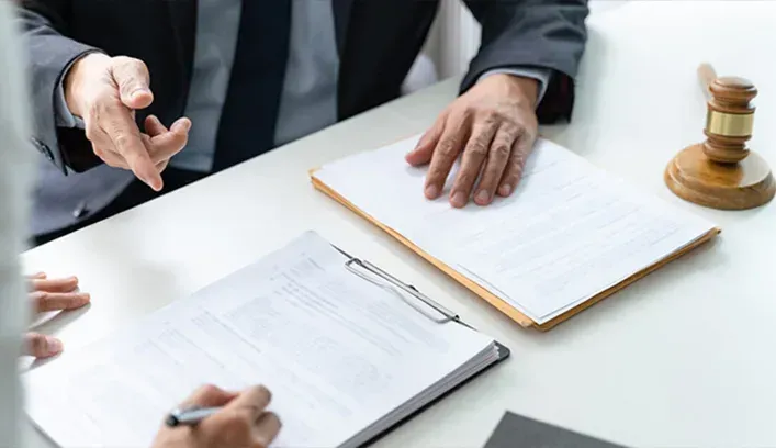 A person in a suit points to a document while another person signs a paper at a desk with a wooden judge's gavel.