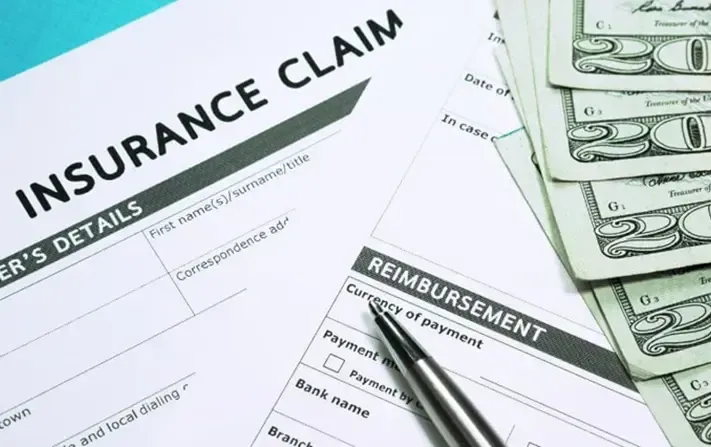 An insurance claim form with a pen resting on it and a stack of twenty-dollar bills next to it.