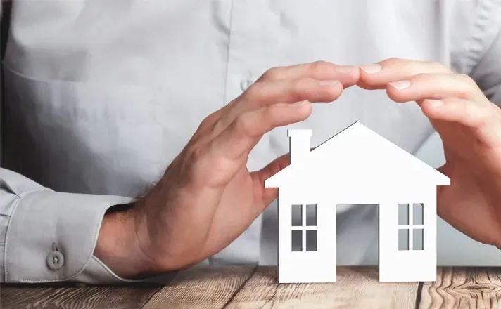 A pair of hands gently cupping and protecting a small, white model house on a wooden surface.