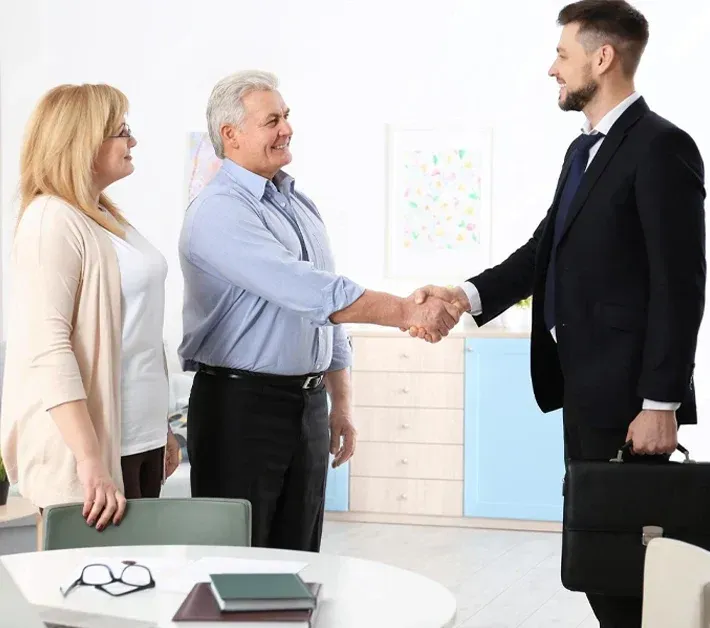 A professional in a suit shakes hands with a person while another individual stands nearby in a bright, modern office.