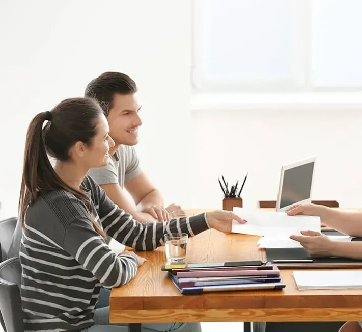 A person in a striped sweater hands a document to another across a wooden desk in a brightly lit office setting.