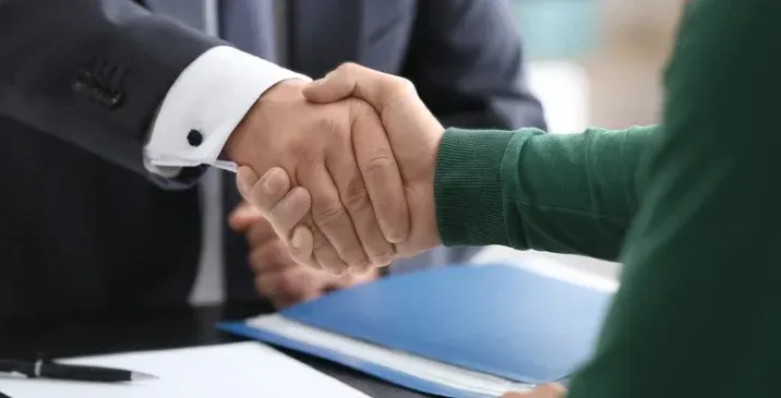 Two people in business attire shaking hands over a desk with a blue folder and a pen.