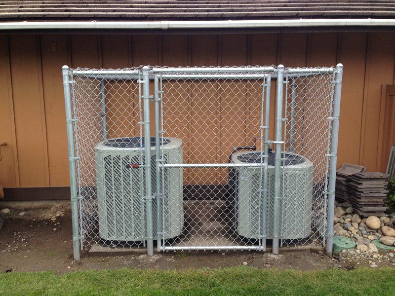 Two air conditioning units enclosed by a chain-link fence, next to a brown building.