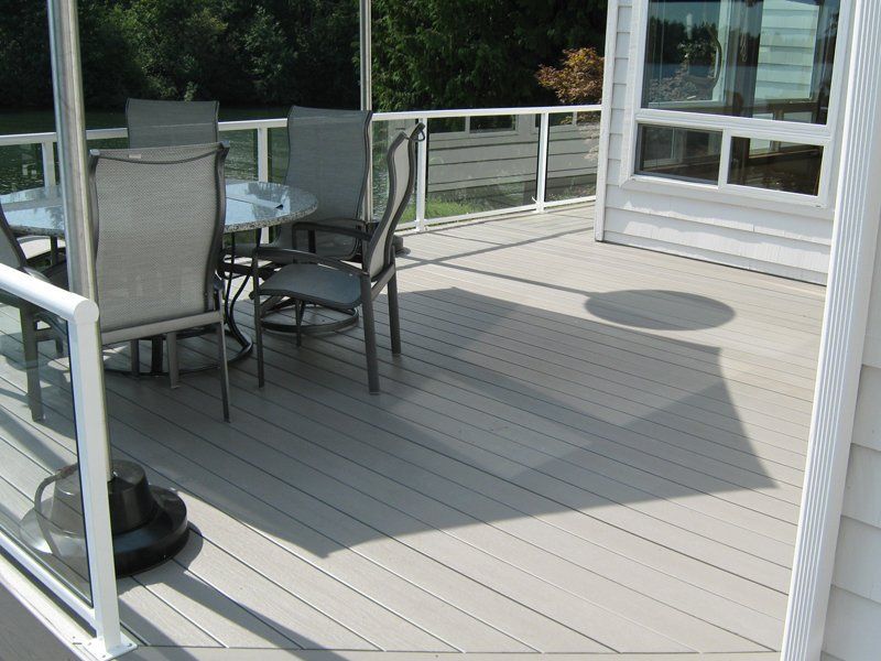 Deck with patio furniture. Grey composite decking, glass railing, table, chairs. Shadow of an umbrella.