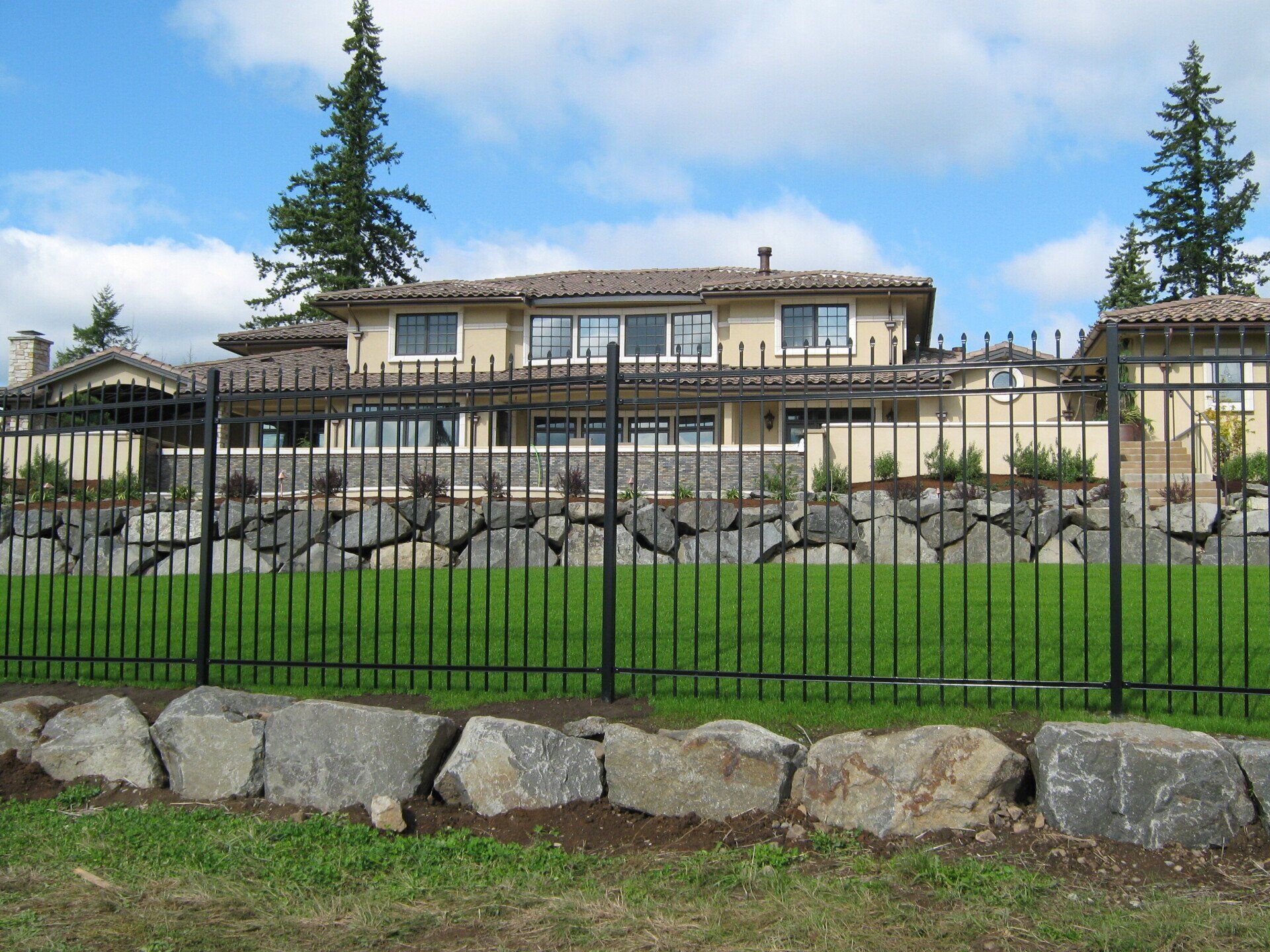 Large beige house behind a black metal fence and stone wall on a grassy lawn under a blue sky.
