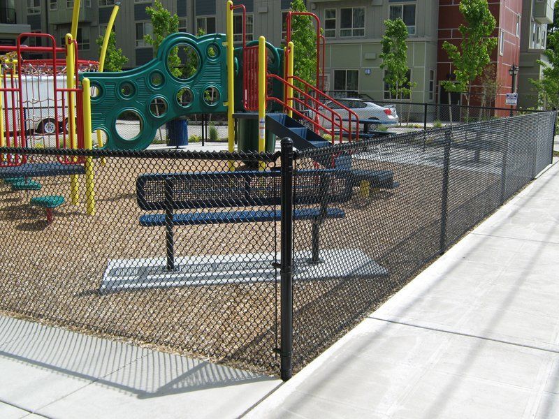 Black chain-link fence surrounds a playground with equipment. Brown wood chips cover the ground. Concrete path visible.