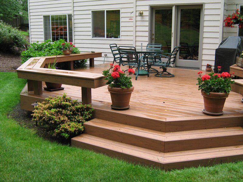Wooden deck with built-in bench, steps, and potted flowers. Patio furniture sits in the background near sliding glass doors.