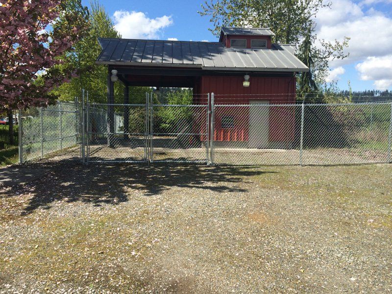 Red building with metal roof, enclosed by a chain link fence. Gravel ground, trees in the background.