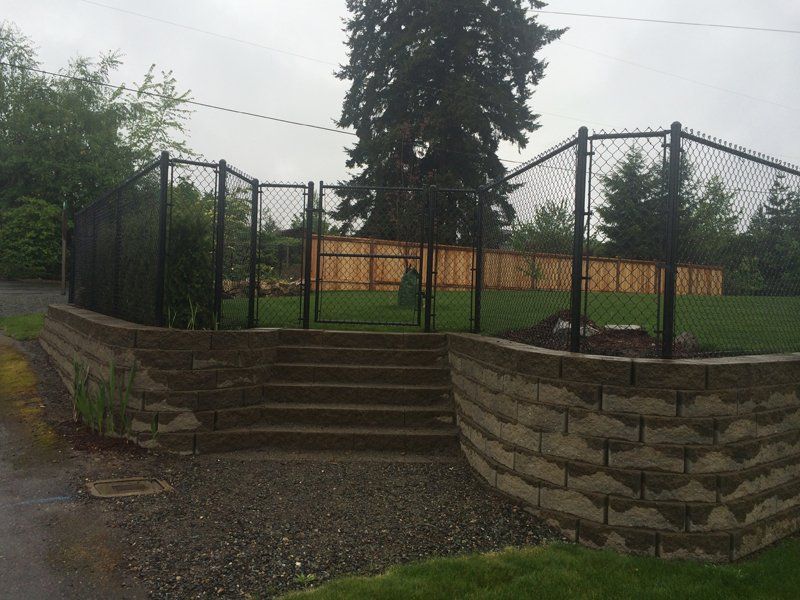 Black chain link fence encloses a green lawn. Brick steps lead up to the gate, set in front of a wood fence.