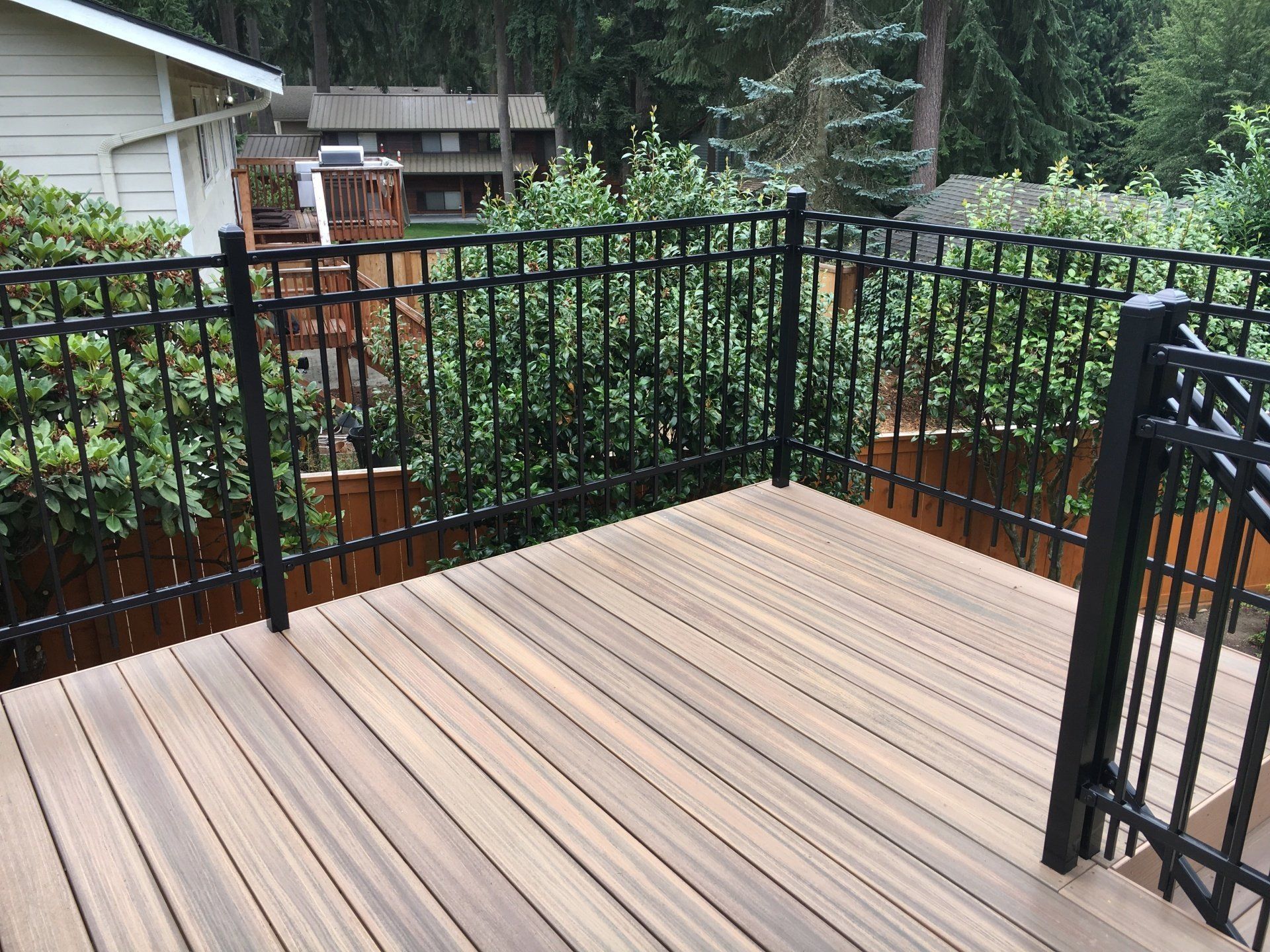 Wooden deck with black metal railing overlooking greenery and a house.