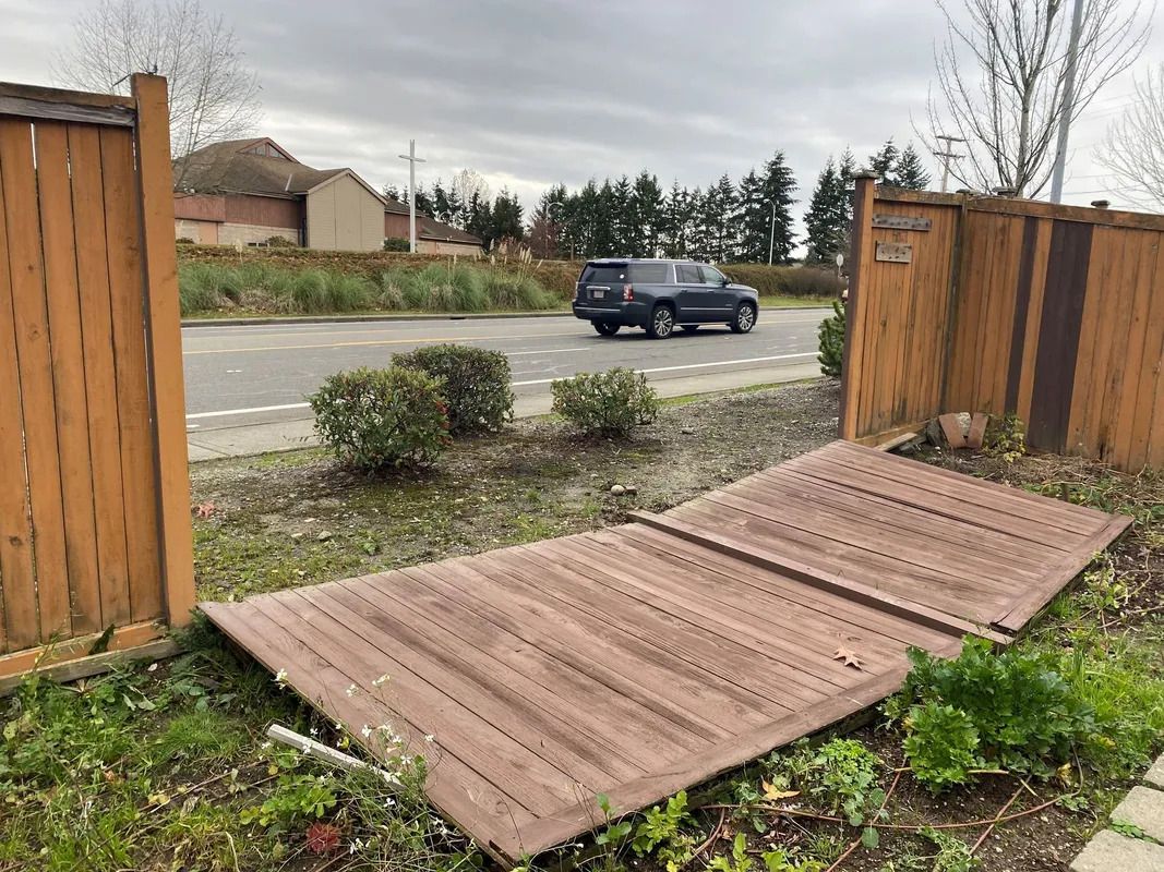 Wooden fence panels, one fallen, next to a road with a car. Cloudy day, green foliage.
