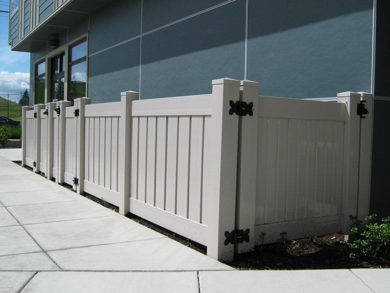 Beige vinyl privacy fence against a building; black hinges and posts.