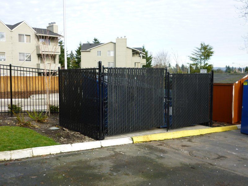 Black fence enclosure for dumpsters in a parking lot, with apartment buildings in the background.