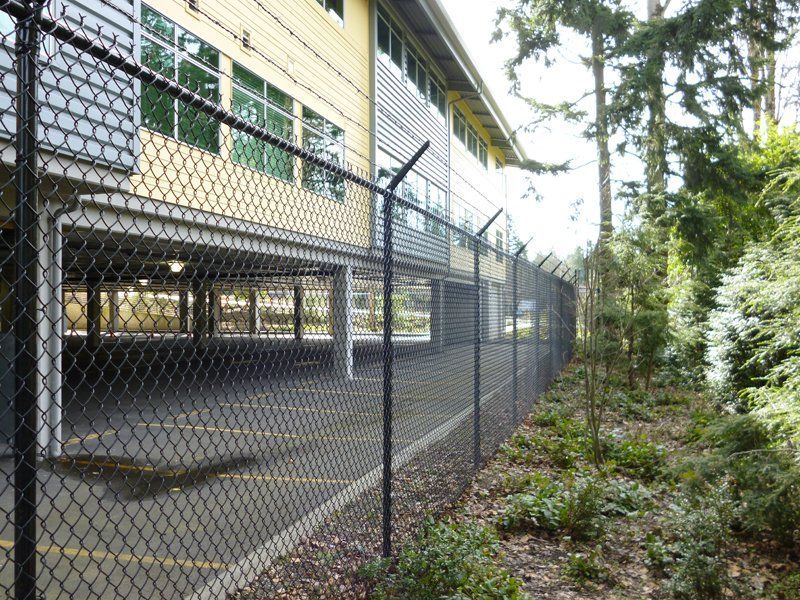 Black chain-link fence with barbed wire surrounds a building with an open parking structure. Trees are visible.