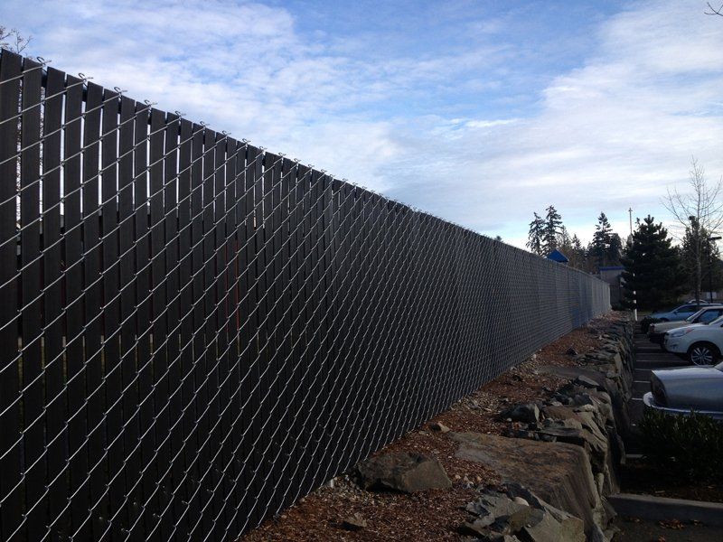 Chain link fence with black slats, alongside a rock retaining wall, with parked cars visible.