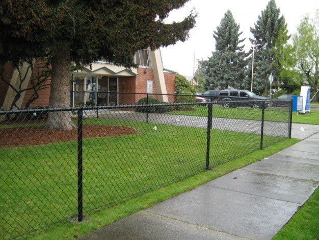 Black chain-link fence bordering a grassy area next to a sidewalk, trees, and a brick building.
