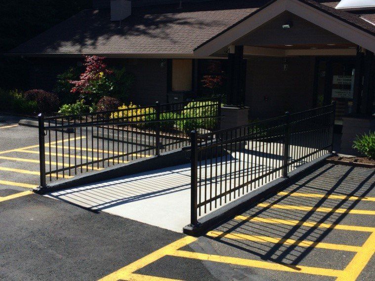 Black metal ramp with railings leading to building entrance, set in asphalt with yellow parking lines.