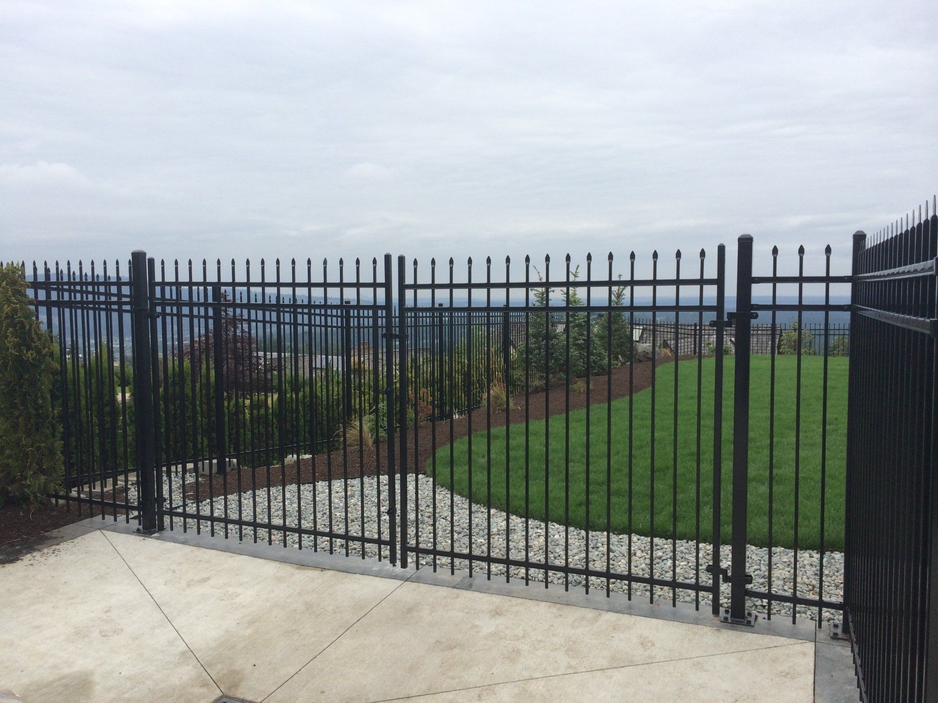 Black metal fence encloses a green lawn, garden, and patio overlooking a cloudy vista.