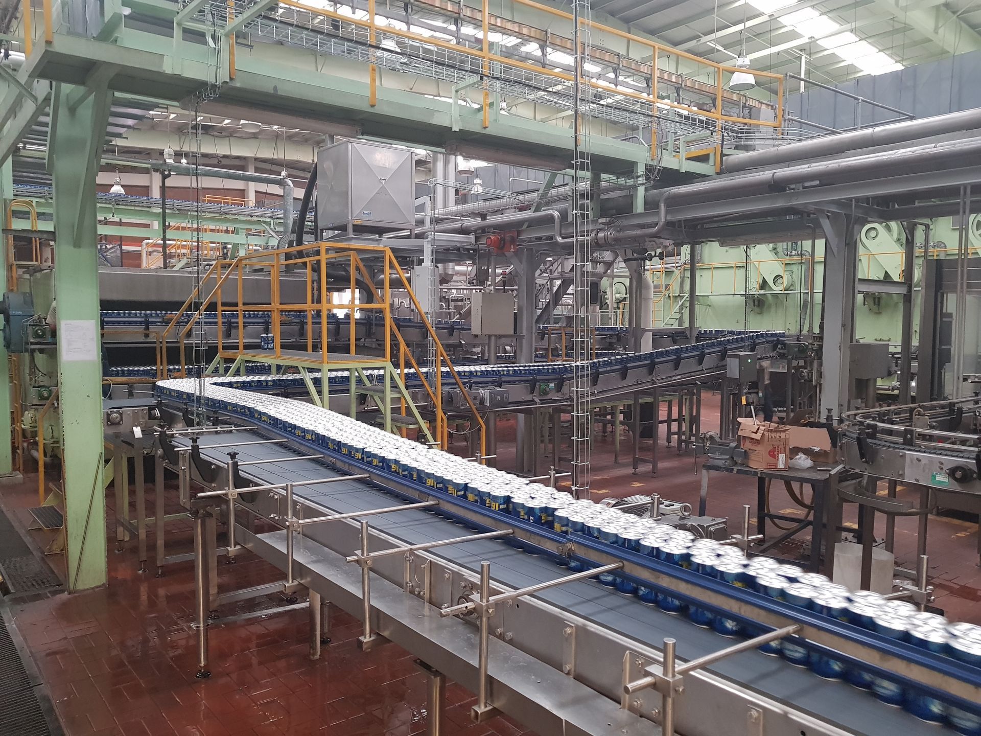 Línea de producción industrial con cintas transportadoras que llevan paquetes blancos bajo tuberías metálicas y andamios.