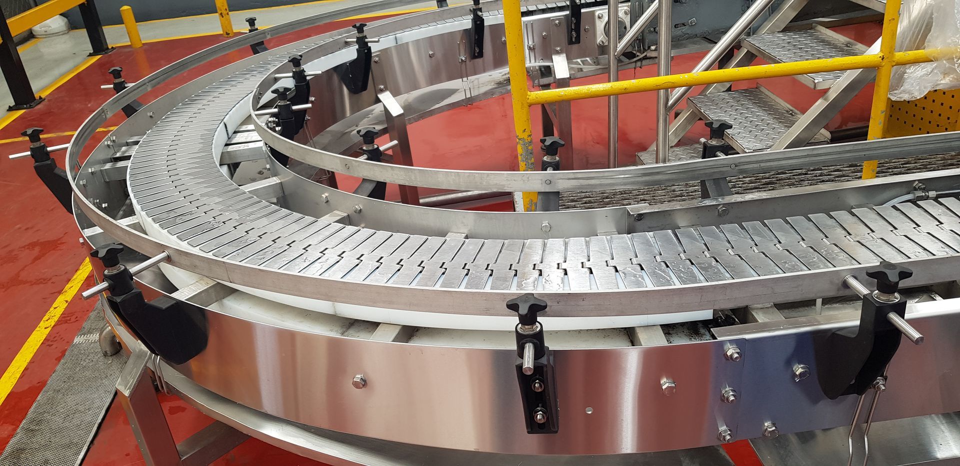 Cinta transportadora metálica curvada en una fábrica industrial con suelo naranja y barandillas de seguridad amarillas.