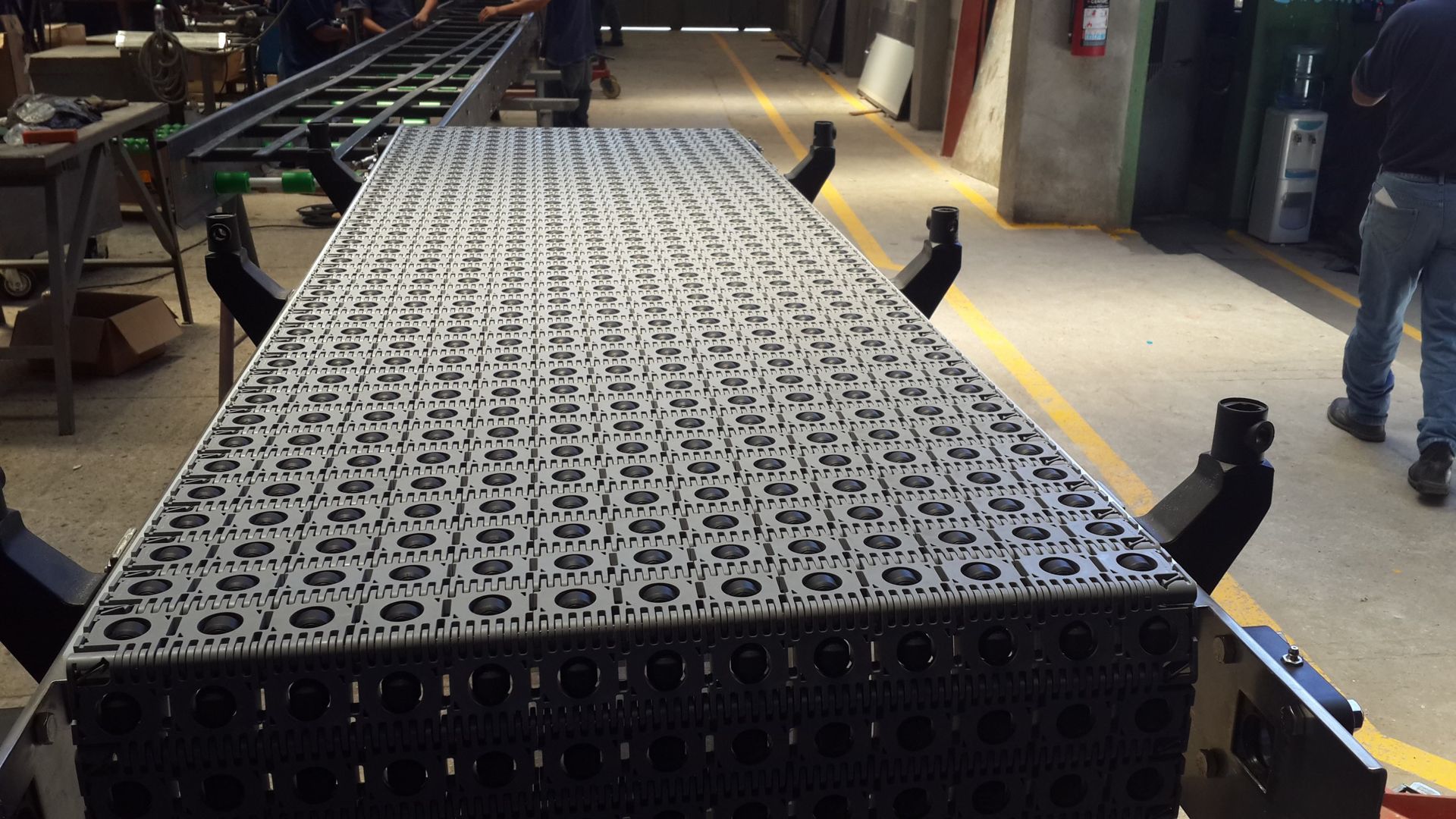 Cajas de plástico negro apiladas en una cinta transportadora de una fábrica, con trabajadores visibles al fondo.