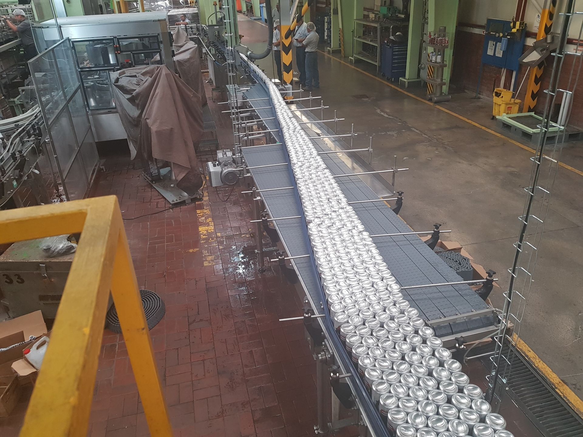 Línea transportadora industrial con componentes metálicos en una fábrica, suelo mojado y trabajadores al fondo.