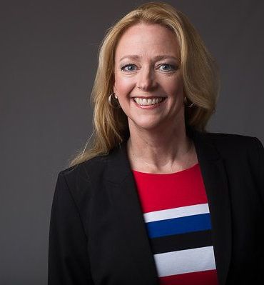 A woman wearing a black jacket and a red and blue striped dress is smiling.