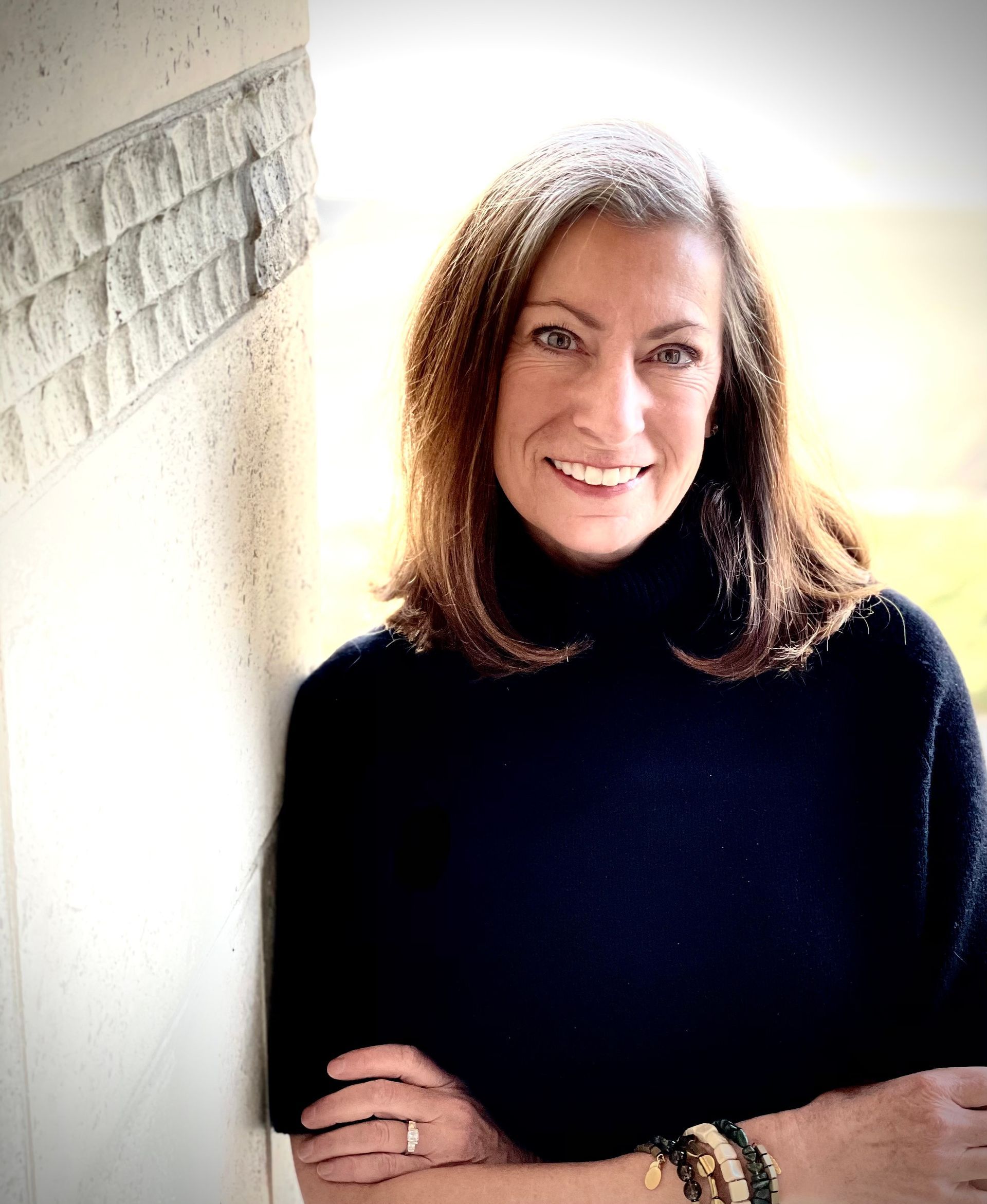 A woman is leaning against a wall with her arms crossed and smiling.