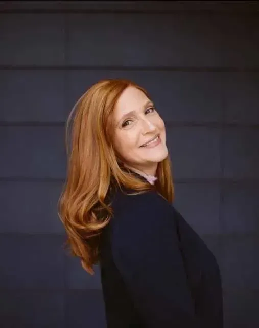 A woman with red hair is wearing a black jacket and smiling.