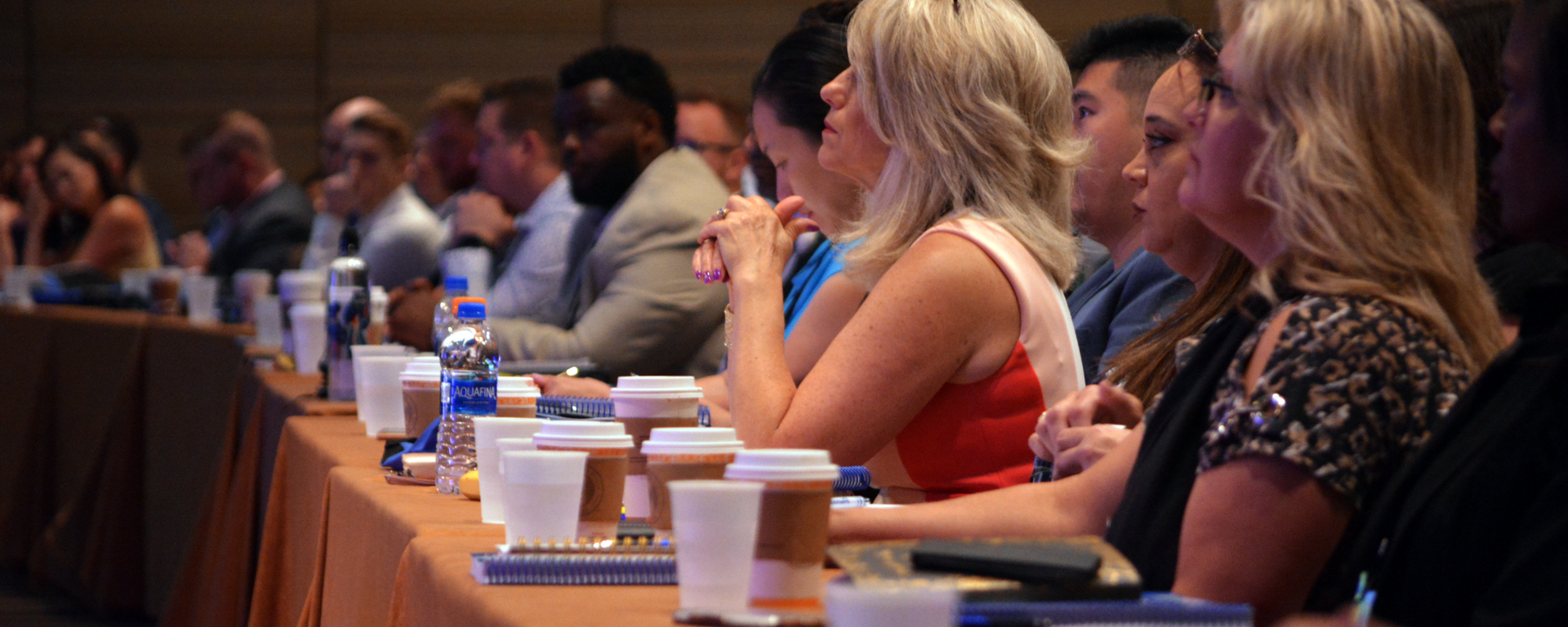 A group of people are sitting at a long table with cups of coffee.