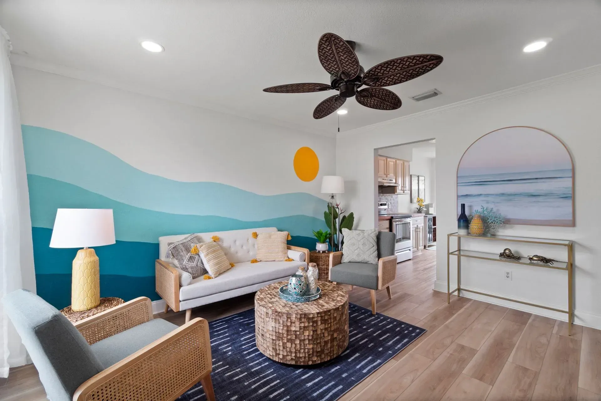 Bright living room with a white sofa, blue wave mural, wicker table, and large ceiling fan