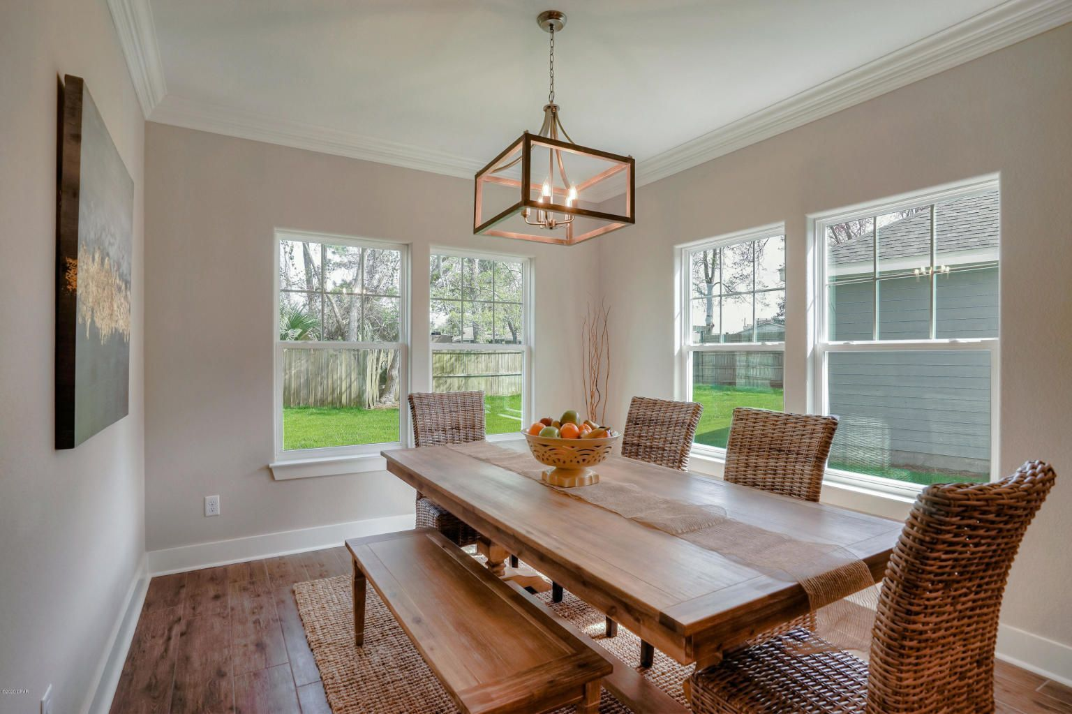 Dining room with wood table, woven chairs, and a bench; three windows overlook a green yard.