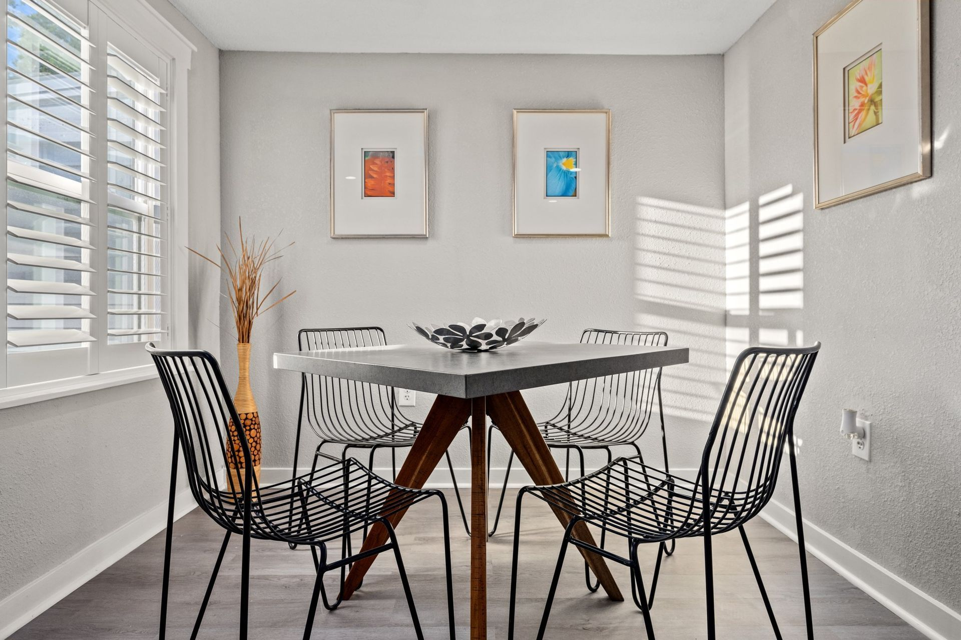 Dining room with square table, black wire chairs, artwork, and shuttered windows.