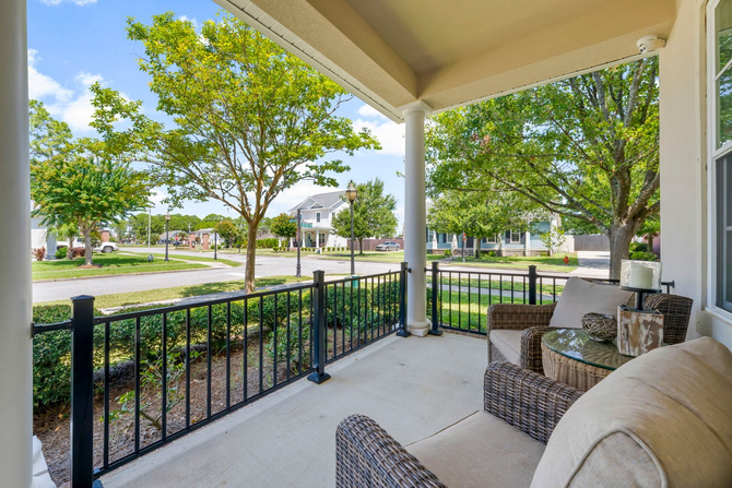Covered porch with seating overlooking a residential street. Green trees, black railing, and blue sky.