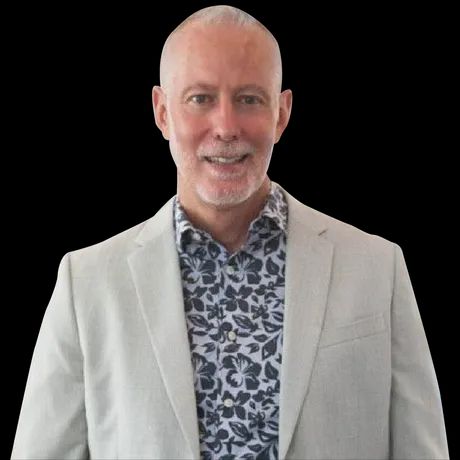 Man in light blazer and floral shirt smiles against black background.