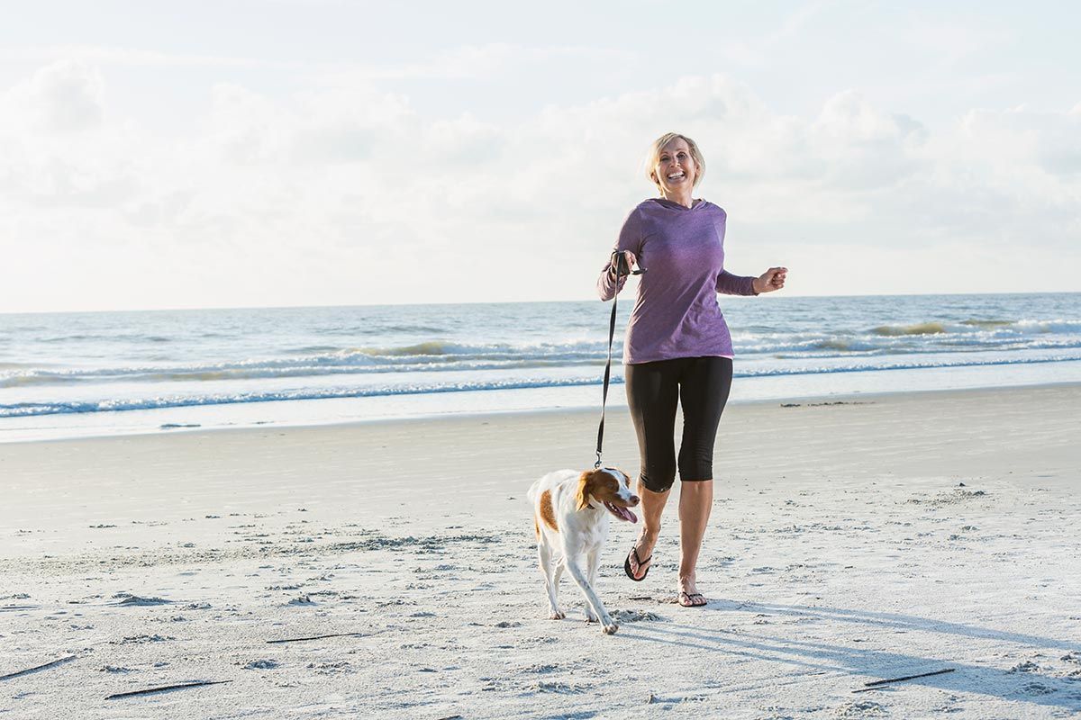 Woman jogging barefoot on a beach with a dog at sunrise