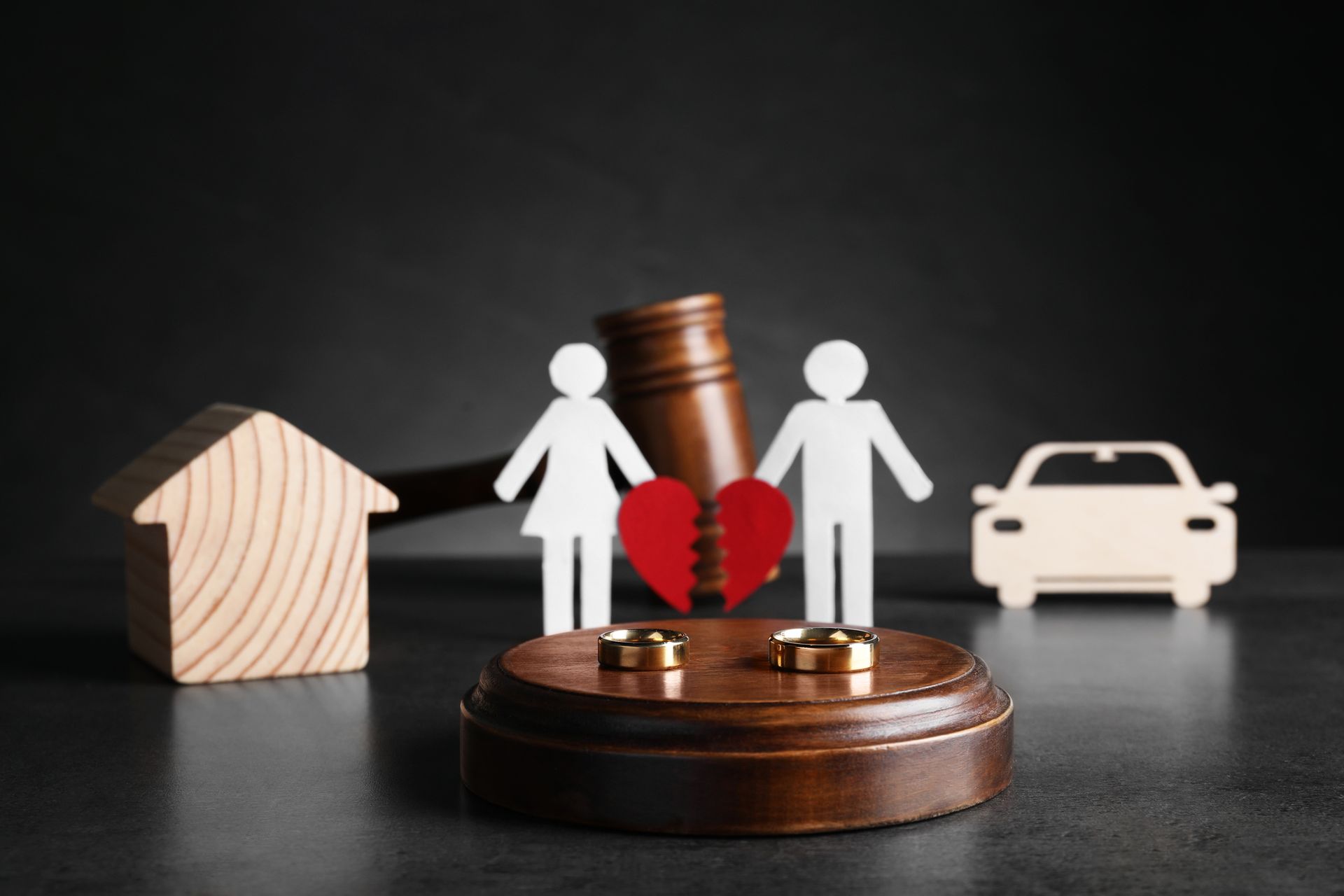 Wedding rings and a paper cutout of a couple with a broken red heart on a grey table.