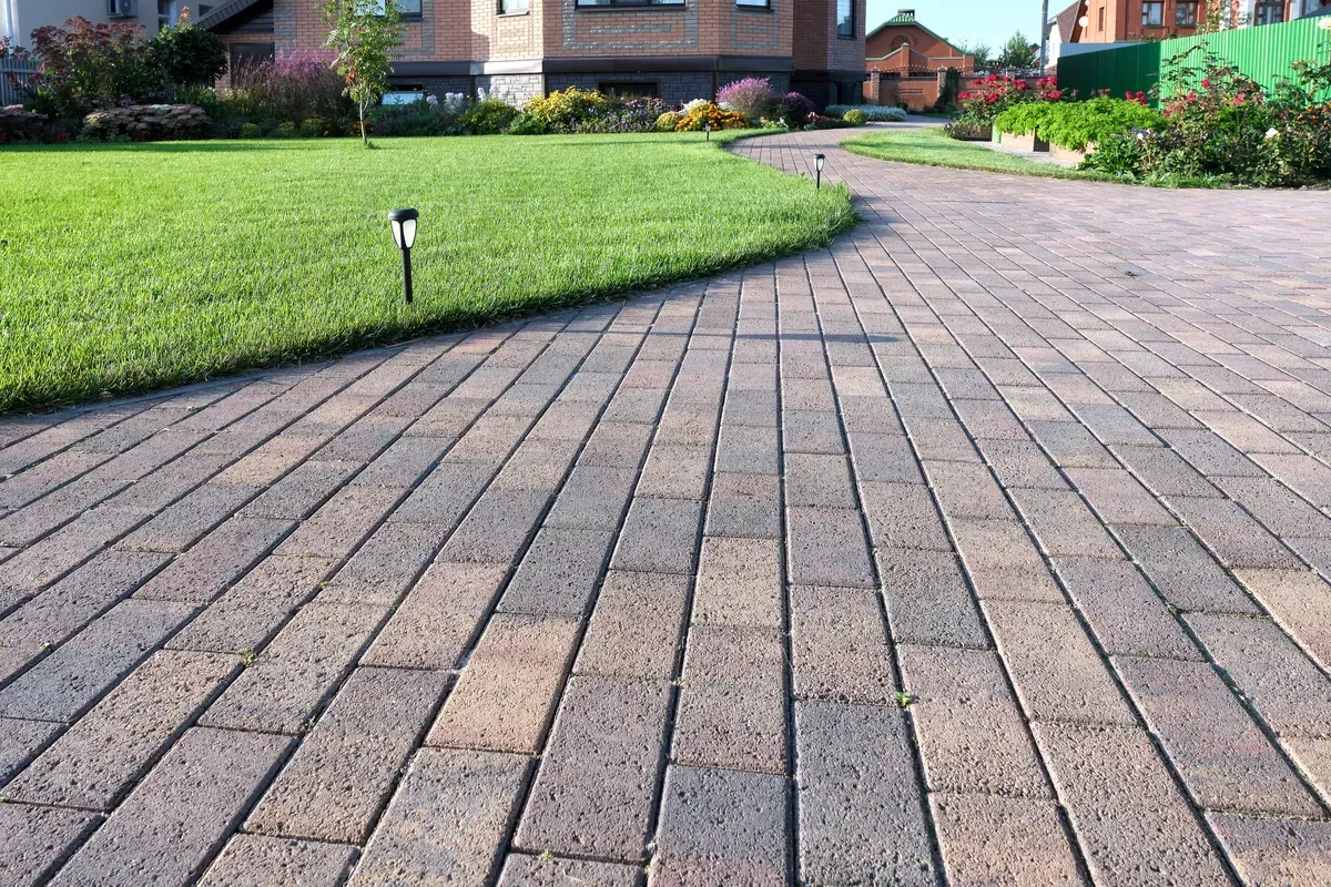 Brick driveway leading to a house, bordered by green lawn and flower beds.