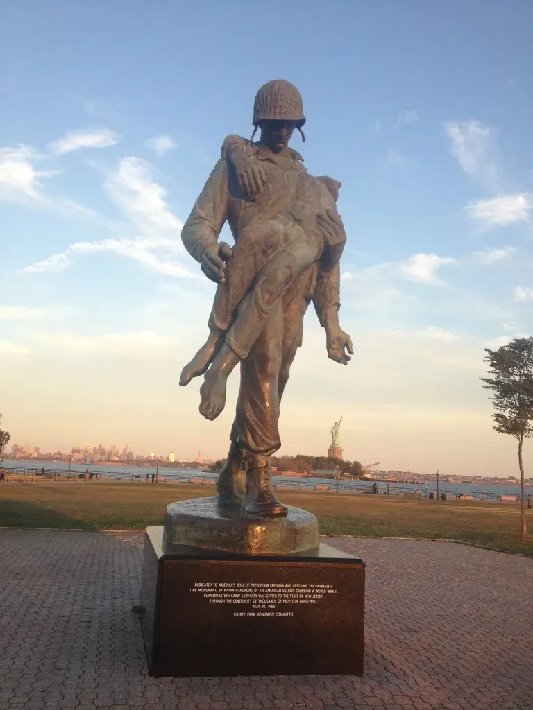 A statue of a soldier carrying a child with the statue of liberty in the background